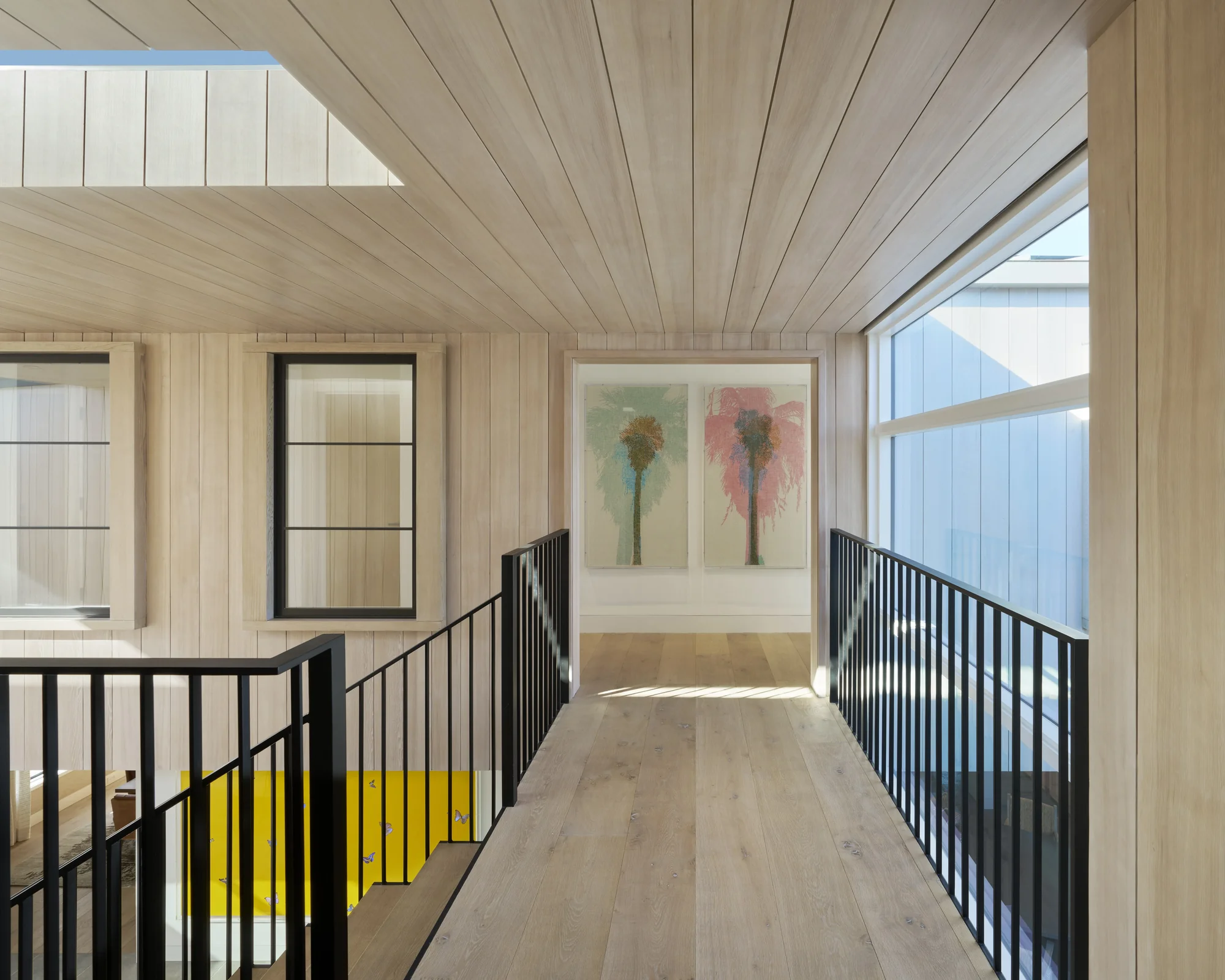 Upper-level hallway with light wood paneling, black metal railings, palm tree artwork, and large windows framing views.