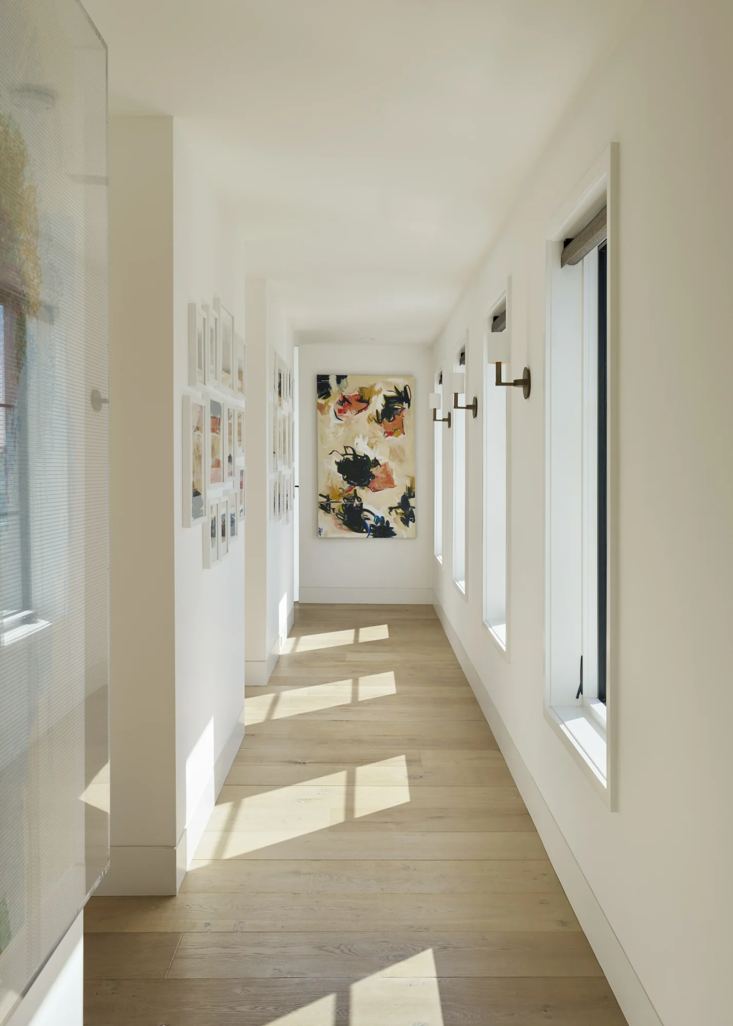 Light-filled hallway with white walls, oak flooring, framed artwork gallery, and colorful abstract painting at corridor's end.