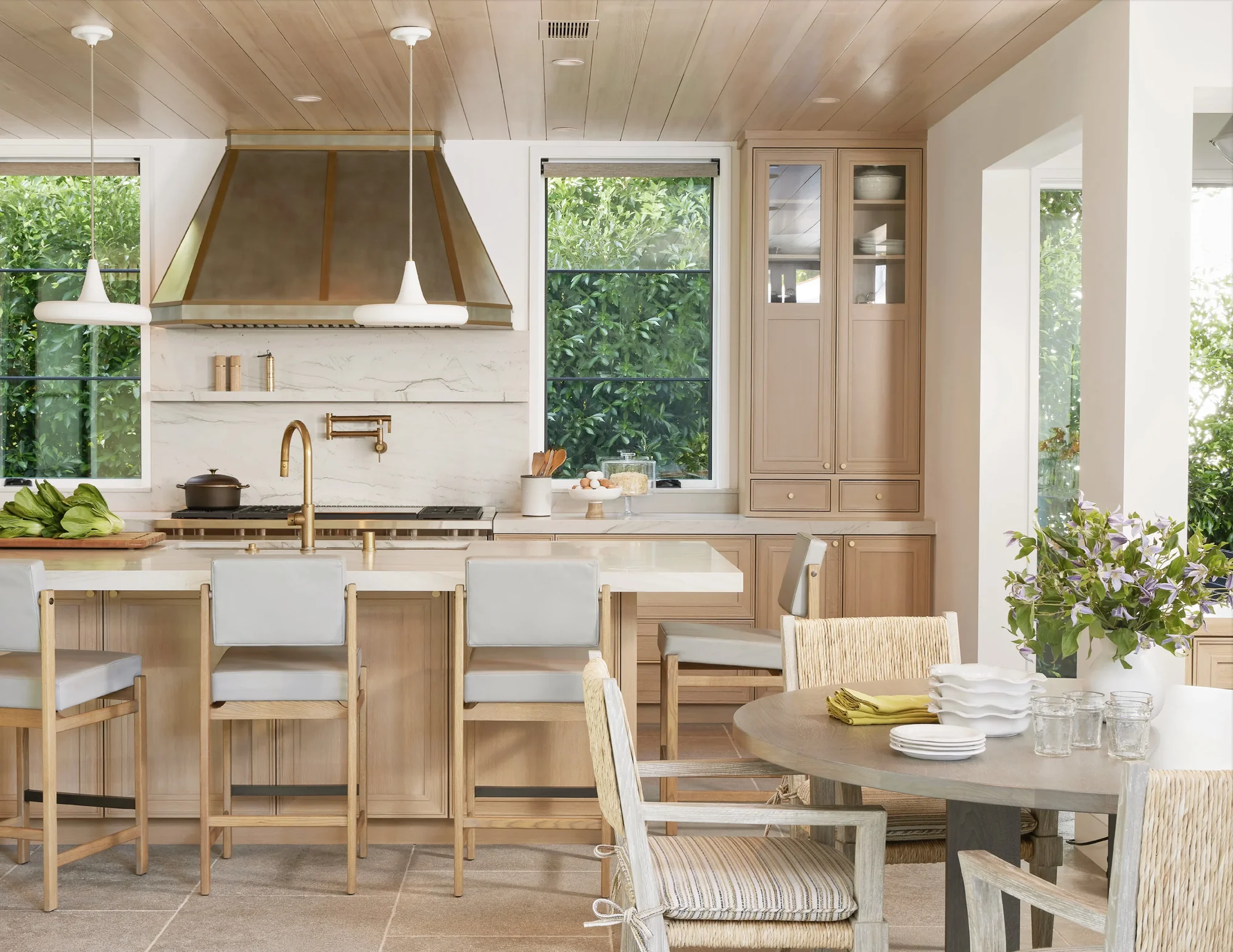 Modern kitchen with natural wood cabinetry, marble countertops, brass fixtures, and dining area with garden views