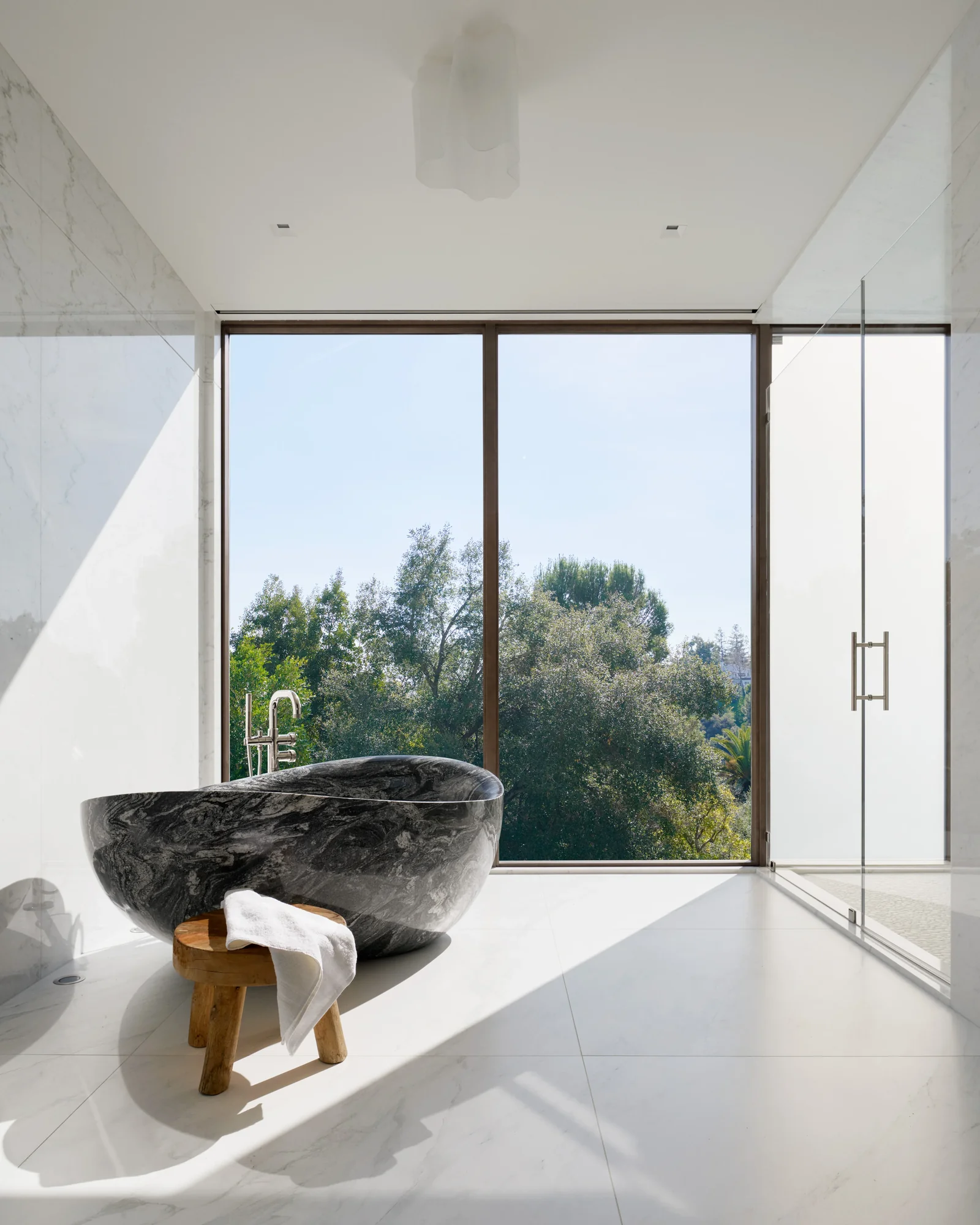 Black marble freestanding soaking tub beside floor-to-ceiling windows overlooking mature trees, with small wood stool and white towel.