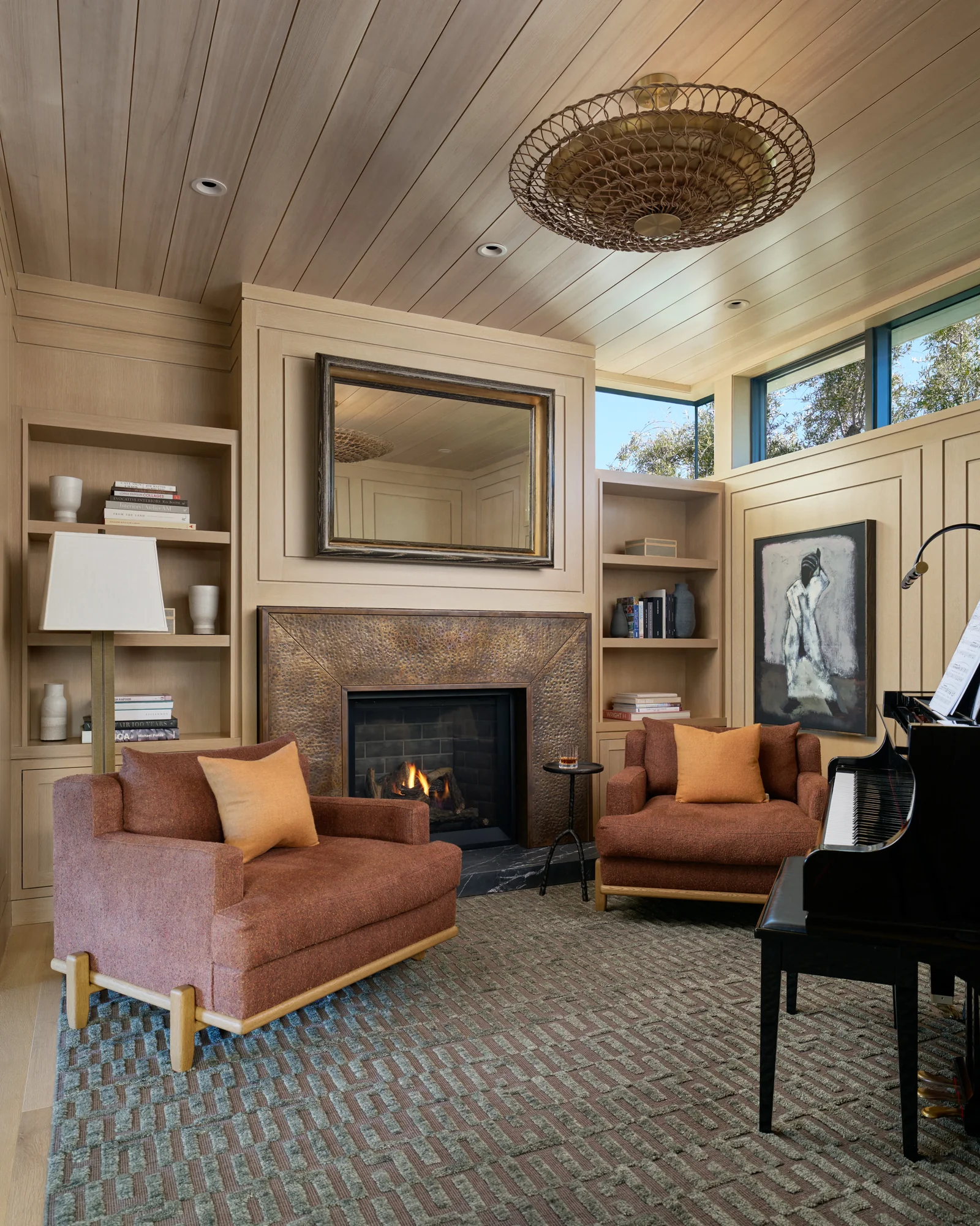 Warm living room with fireplace, built-in shelving, wood ceiling, rust-colored chairs, and piano beneath basket pendant light