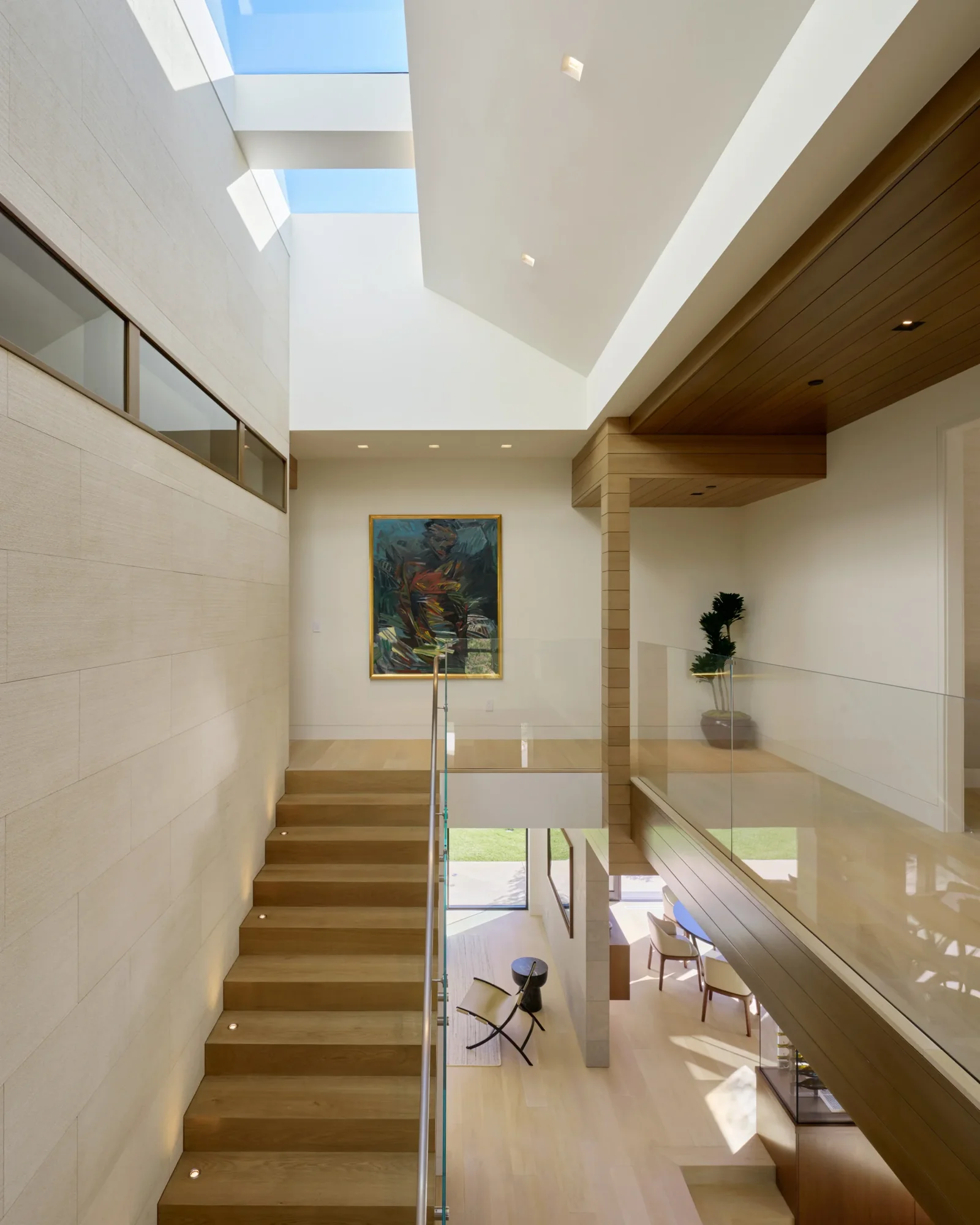 Sun-drenched double-height stairwell with warm wood treads, glass railing, skylight, and open views to outdoor living below.