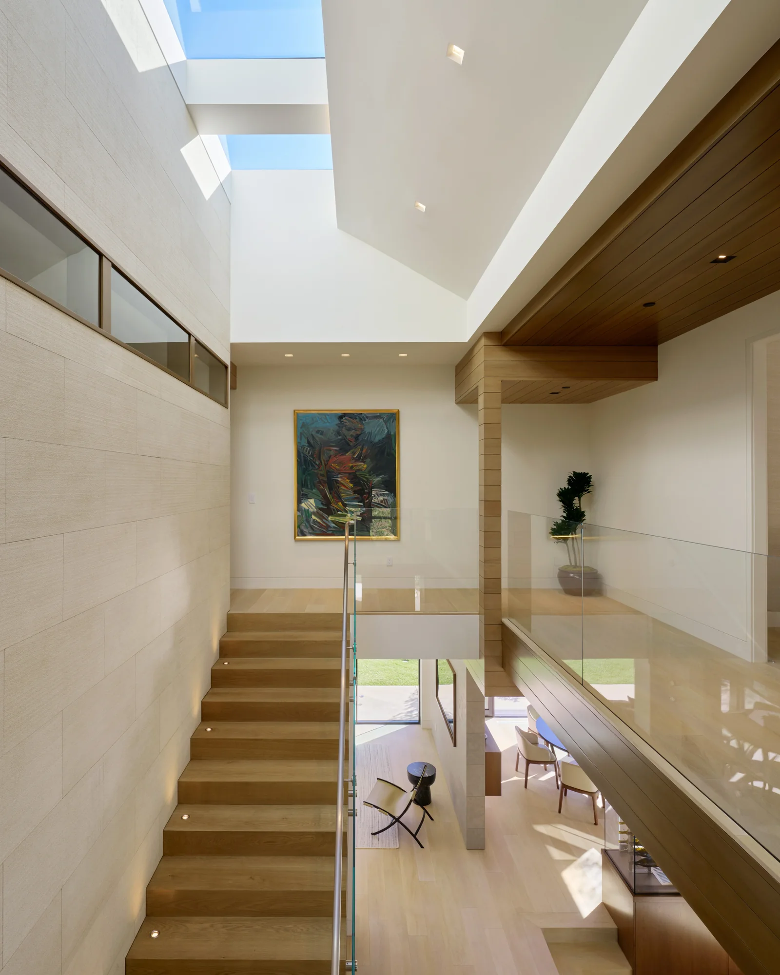 Sun-drenched double-height stairwell with warm wood treads, glass railing, skylight, and open views to outdoor living below.