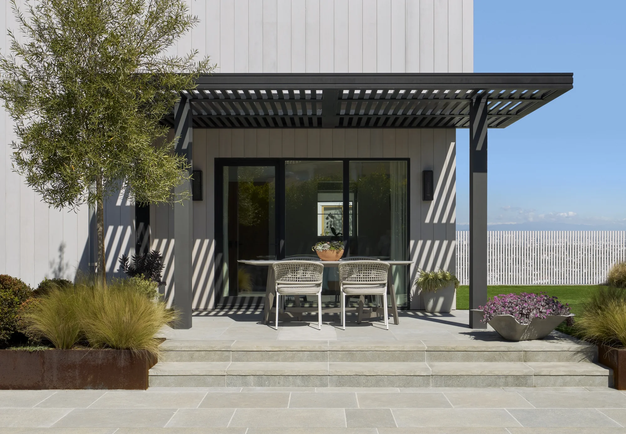 Modern outdoor dining patio with black pergola, glass doors, stone flooring, and desert landscaping under blue sky.
