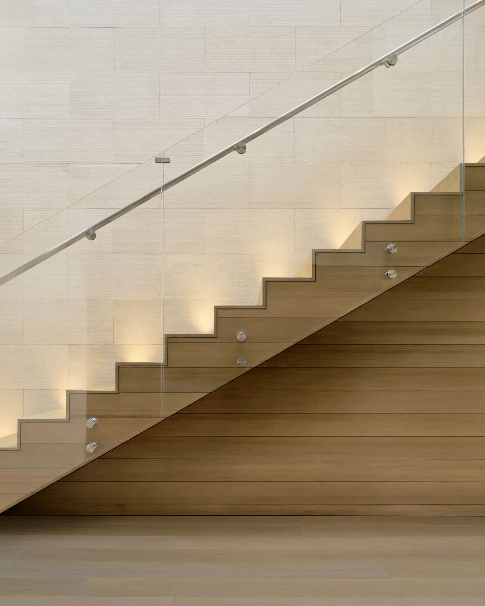 Floating wood staircase with glass balustrade and steel handrail, warm step lighting casting shadows on limestone wall.
