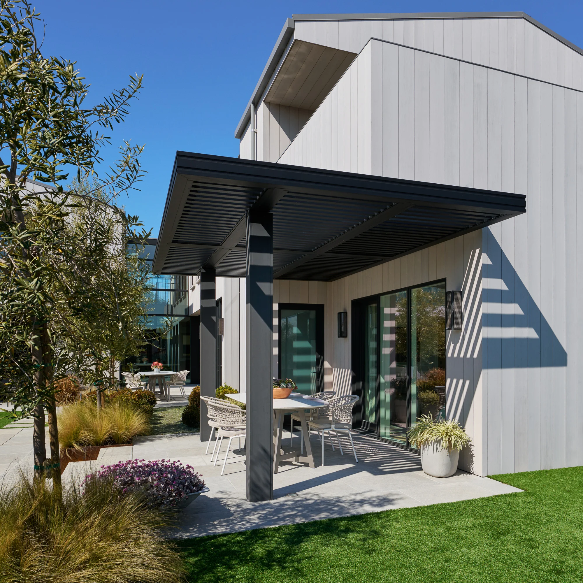 Modern home exterior with black pergola and white outdoor furniture, featuring vertical siding and landscaped patio.