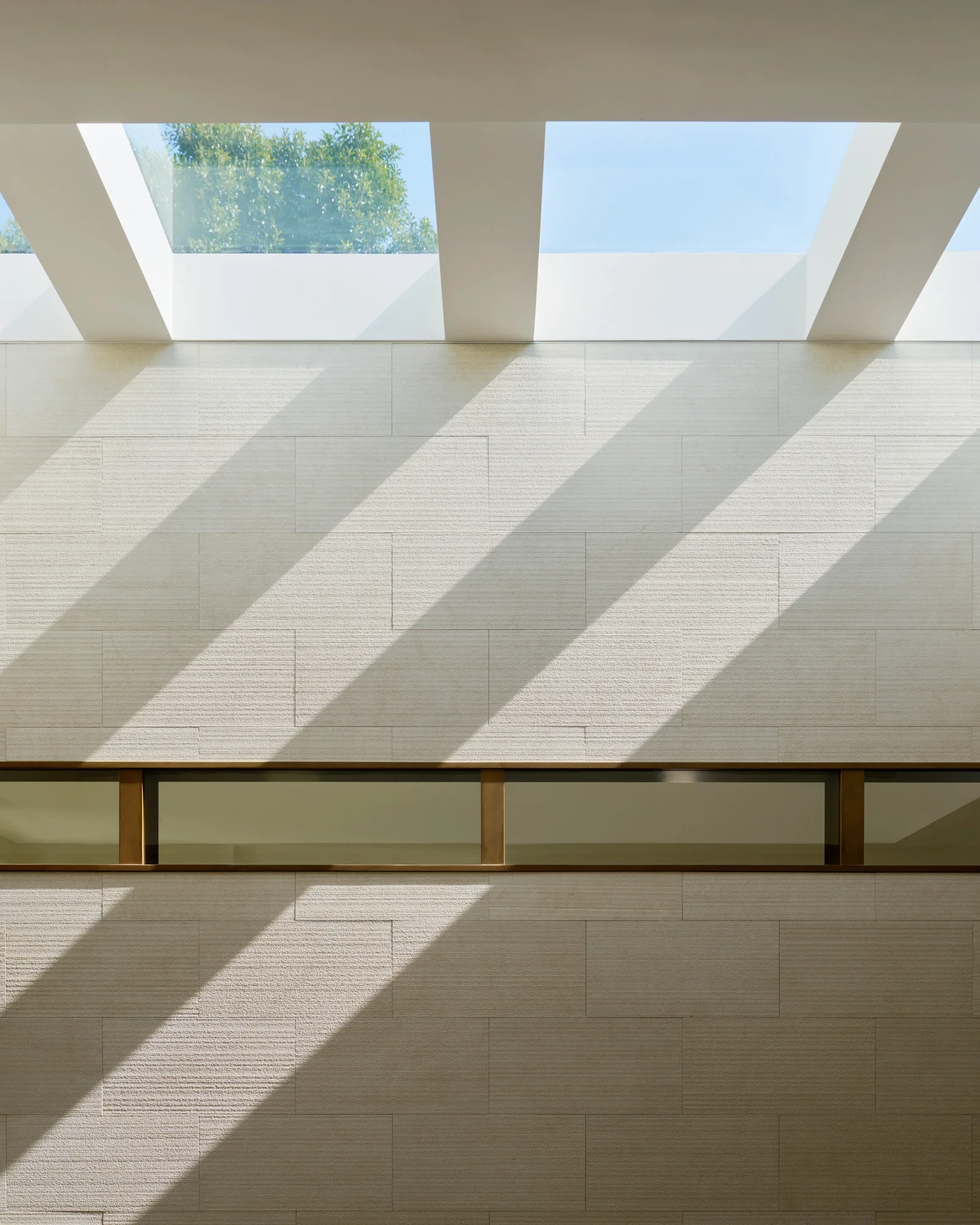 Diagonal light shafts from twin skylights cast dramatic shadows across textured limestone wall panels.
