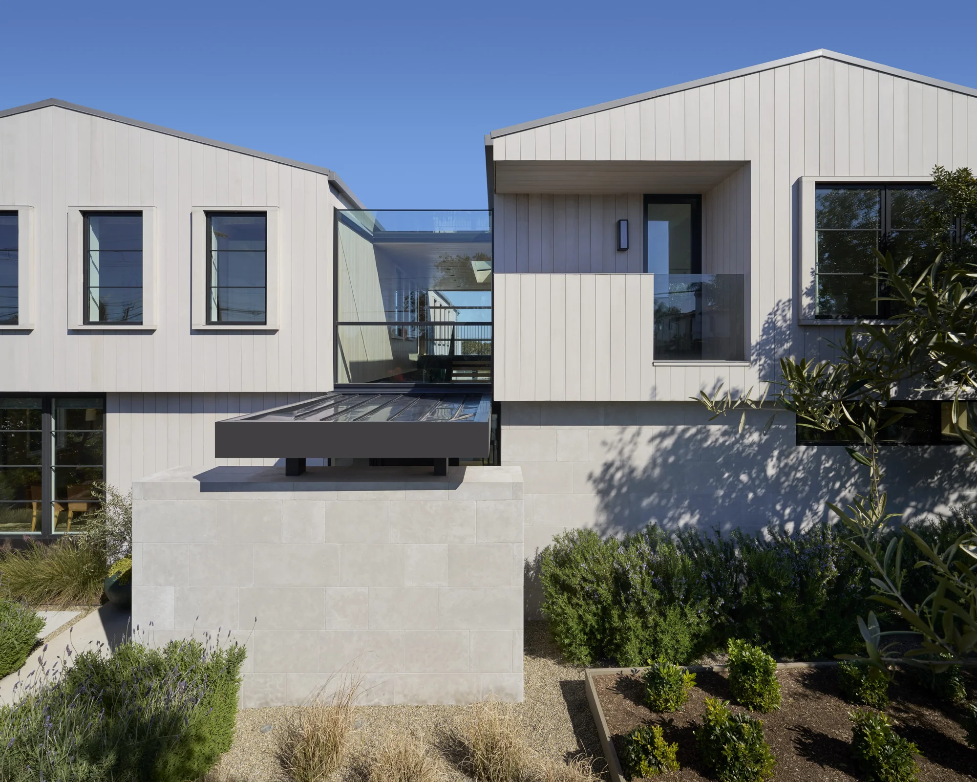 Modern multi-level home with vertical white siding, glass bridge connector, and native landscaping under blue sky.