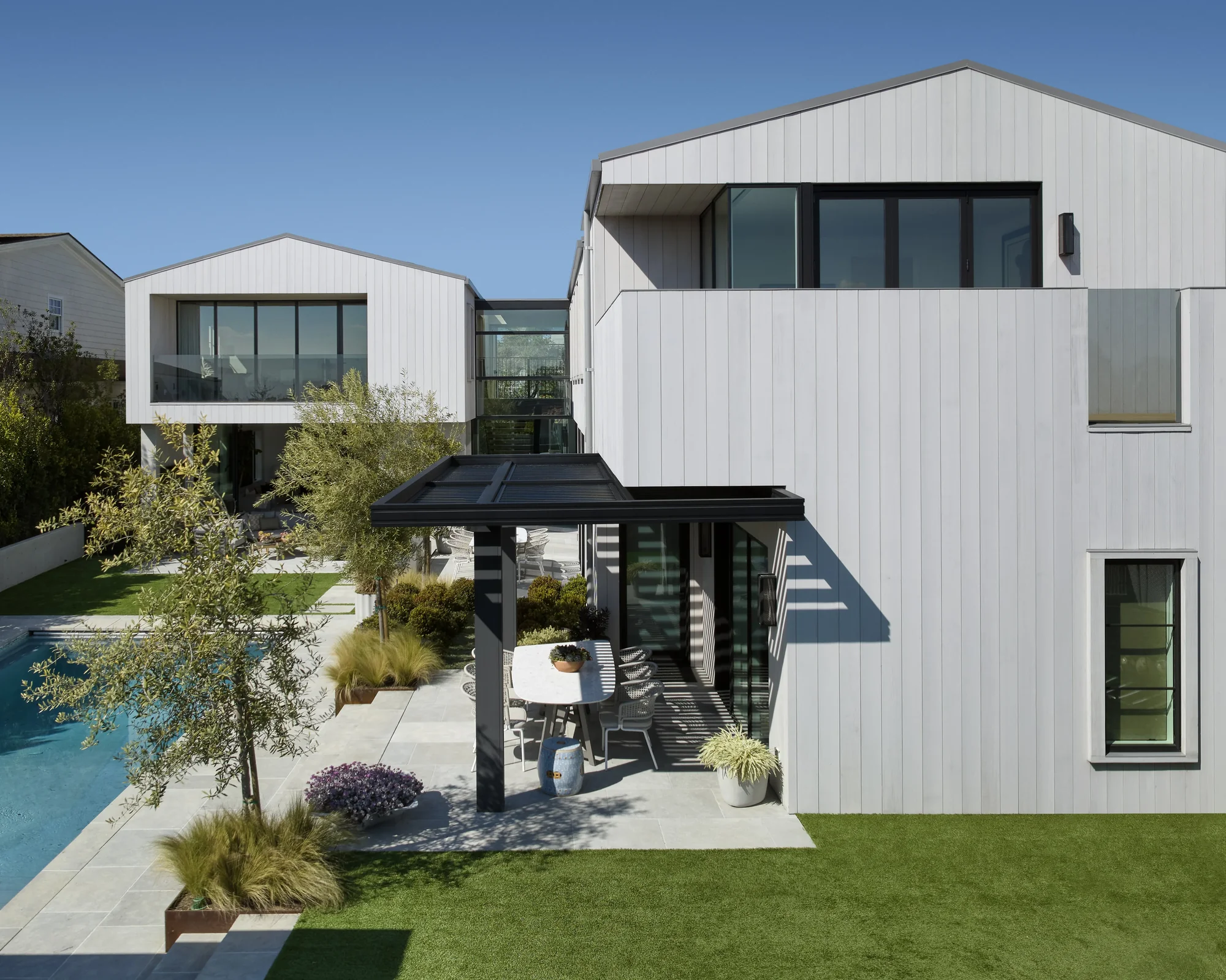 Modern white residential architecture with multiple gabled volumes, outdoor pergola, pool, and landscaped yard under blue sky.