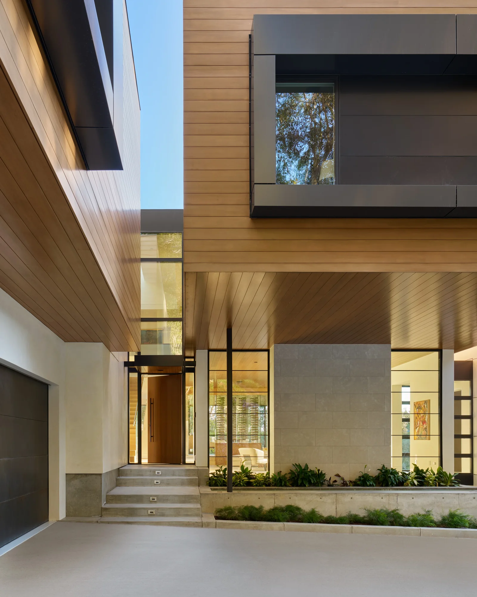 Exterior entry of a modern custom home with cedar siding, stone accents, floor-to-ceiling glass, and a warm wood pivot door.