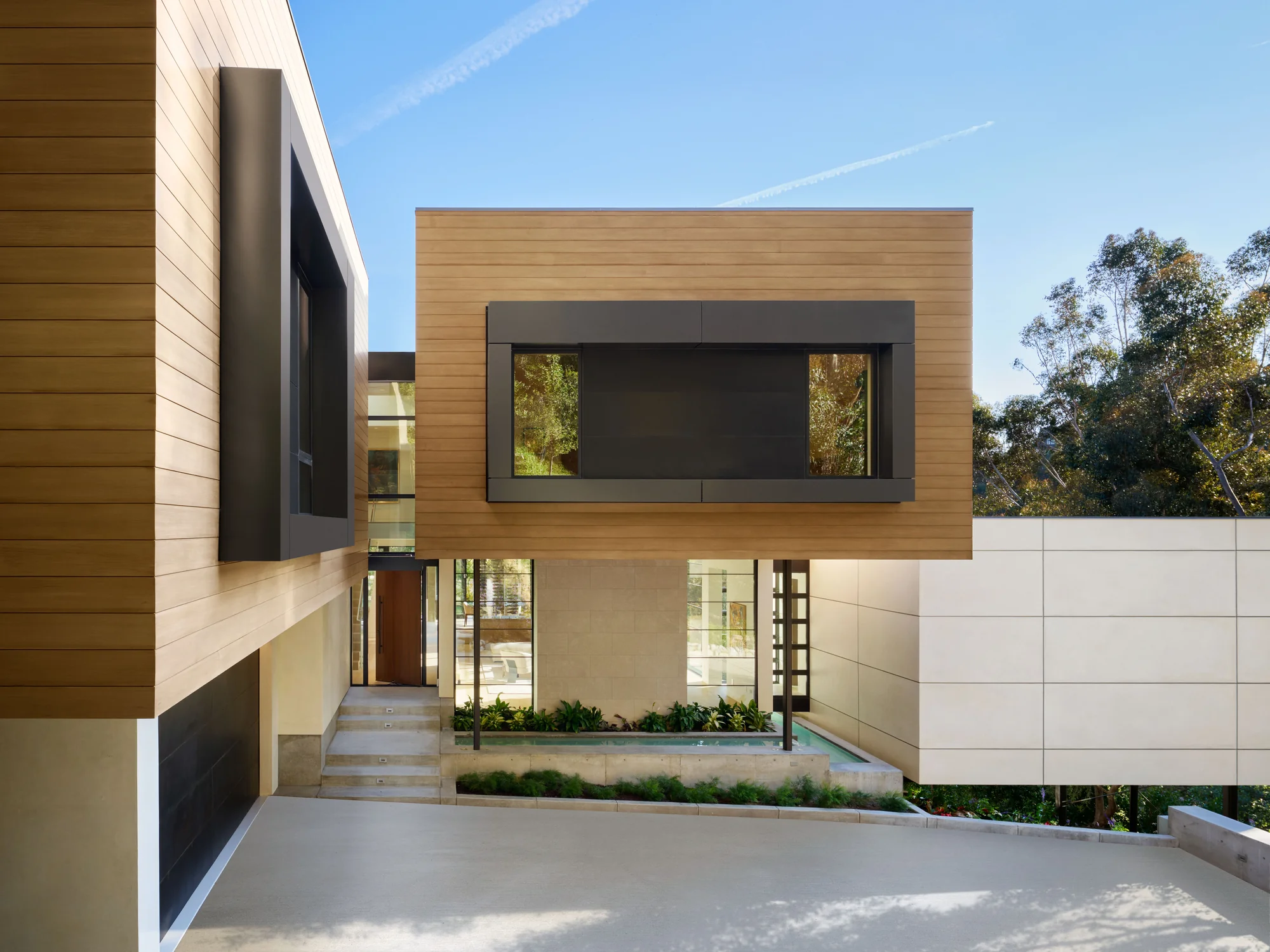 Exterior view of a modern home with cedar siding, dark steel accents, and a cantilevered upper volume above an entry courtyard with a reflecting pool.
