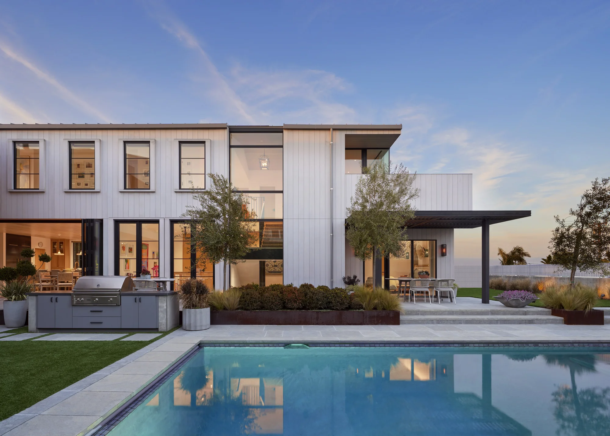 Modern white two-story home with outdoor kitchen and covered patio beside pool at dusk, showcasing indoor-outdoor living.