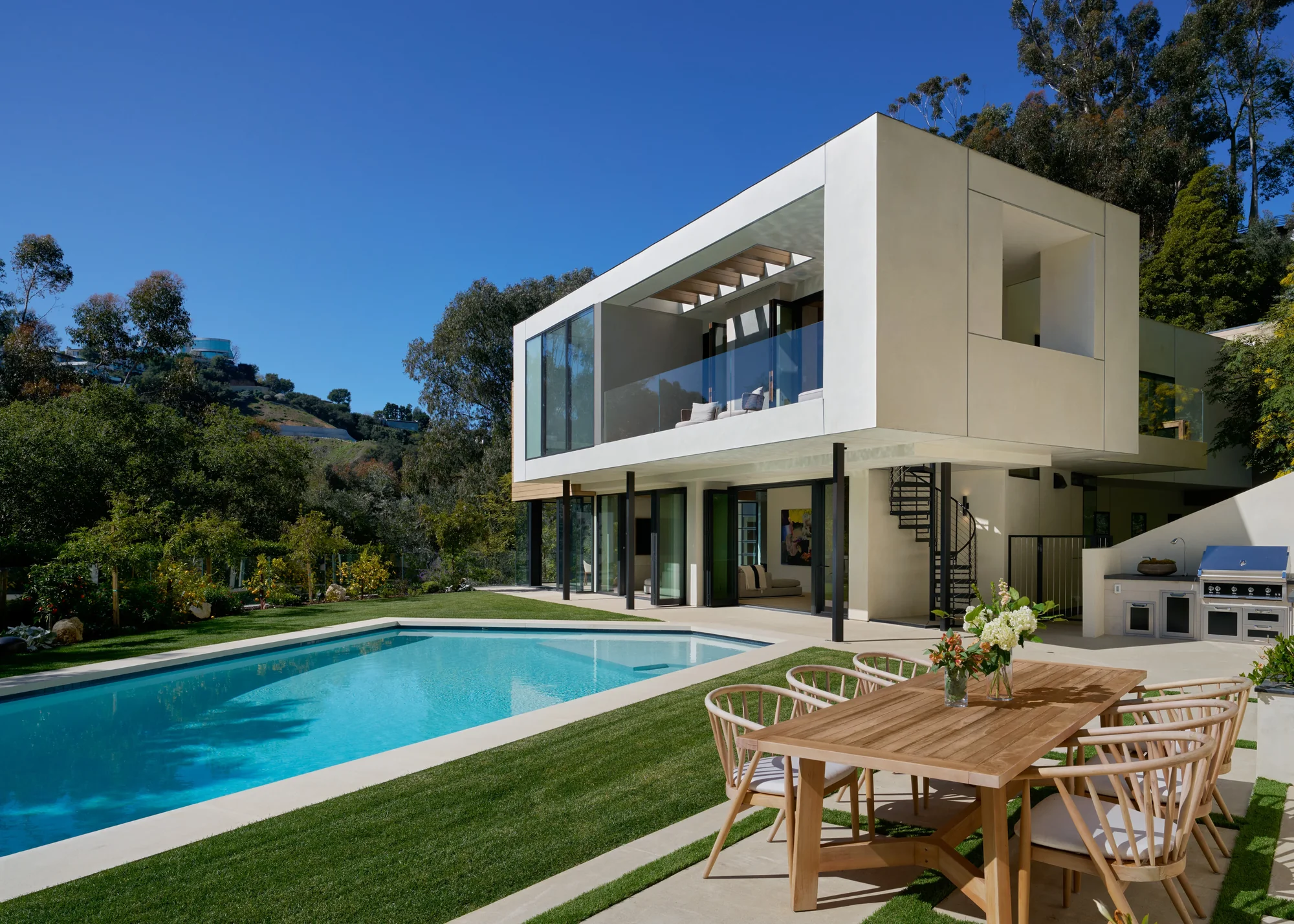 Modern two-story white stucco home with glass walls opens to pool, lawn, and outdoor dining amid hillside trees.