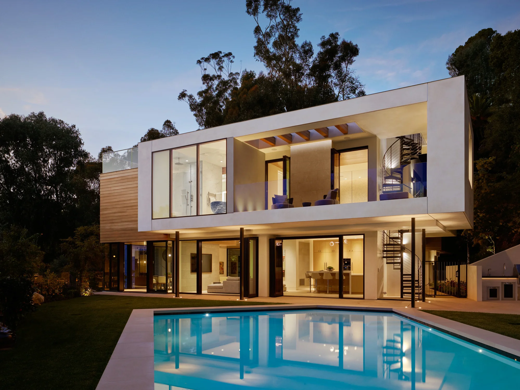 Two-story modern home at dusk with floor-to-ceiling glass, cedar cladding, open interiors, and illuminated pool.